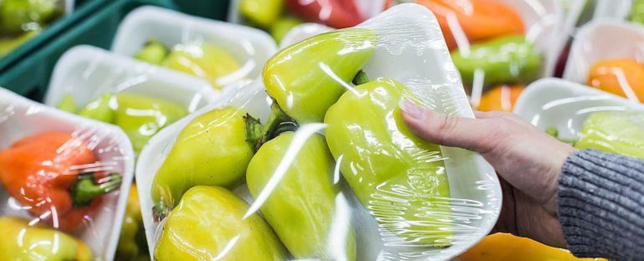 A hand holding a packaged tray of fresh peppers wrapped in plastic, displayed among similar packaging in a grocery store, illustrating the use of single-use plastic packaging in food products.