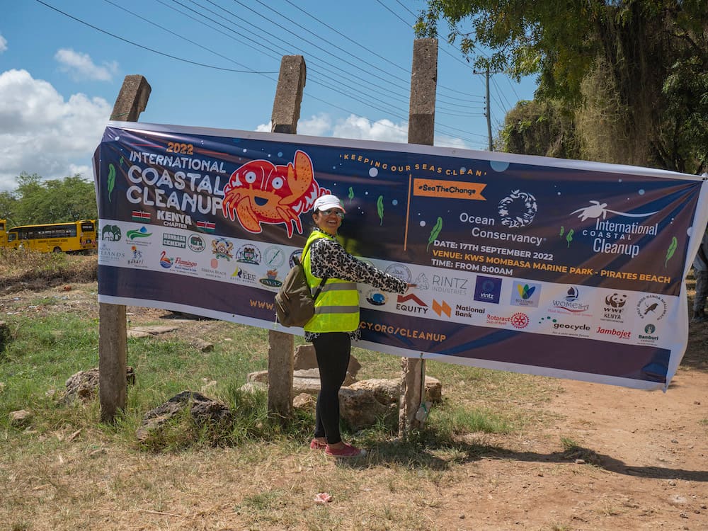 A participant points at a colorful banner for the 2022 International Coastal Cleanup Day in Kenya, featuring sponsors and event details at Pirates Beach in Mombasa.