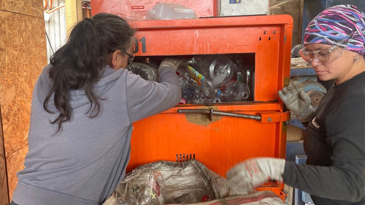 Two women are seen working together at a recycling facility, loading plastic waste into an industrial compactor machine. One wears protective goggles and gloves, emphasizing safety measures in waste management operations. The image represents women's leadership and participation in sustainable waste management.